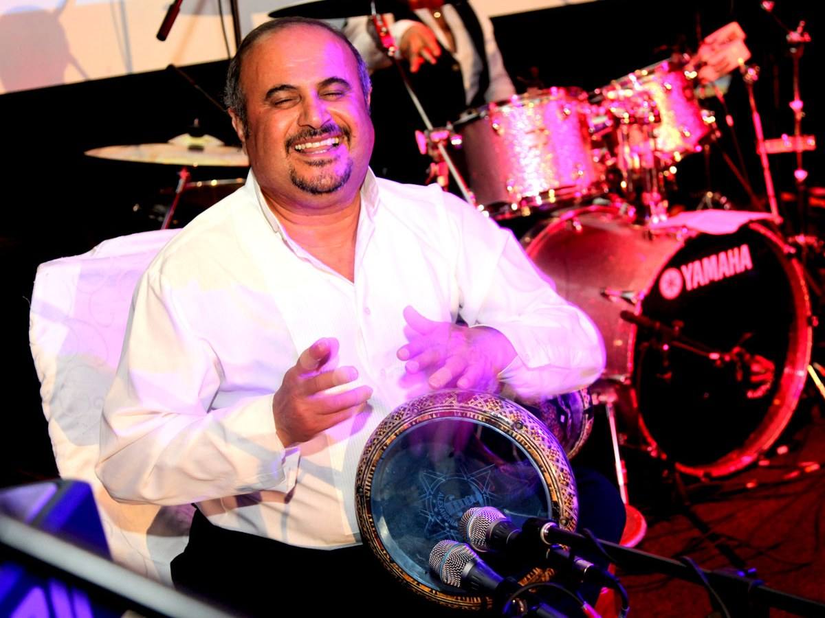 Percussionist playing a hand drum and singing into a stage mic, drum kit behind him